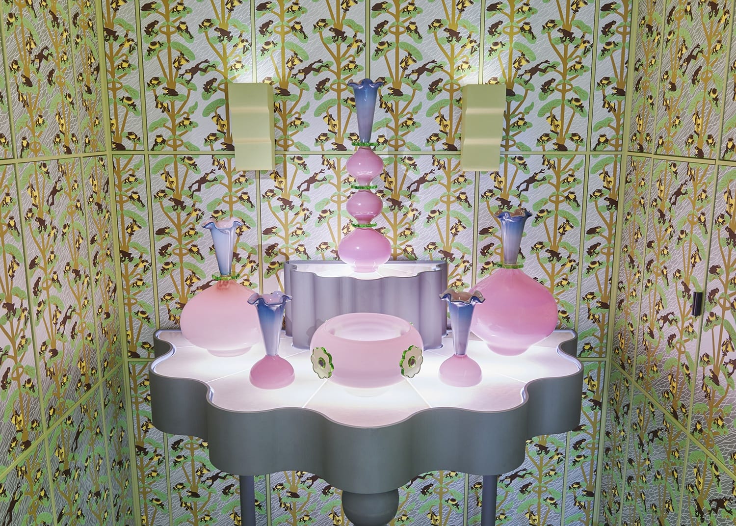 Floral-patterned room with a display of pink and purple glass ornaments set on a wavy table, illuminated by soft lighting.