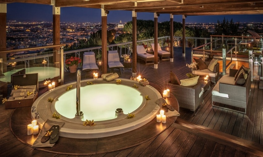 Luxurious rooftop terrace with a glowing hot tub, cozy seating, lit candles, and stunning city skyline views at night.