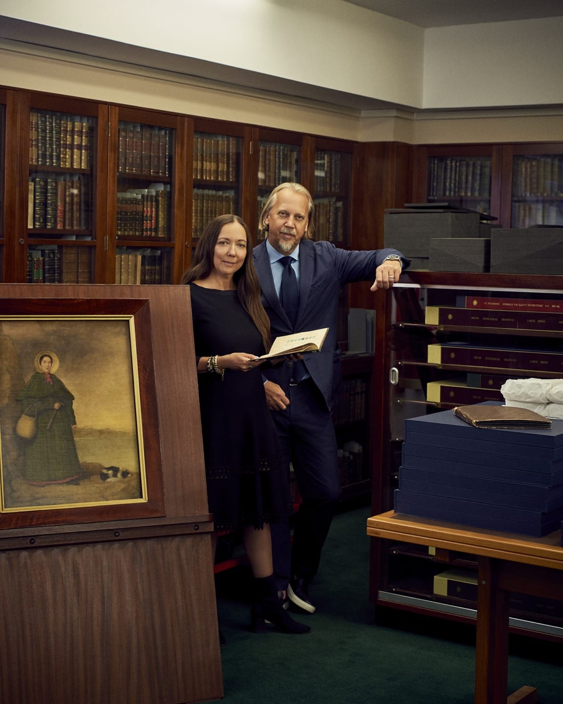 Two people standing in a room with bookshelves, holding a document, with framed artwork and archival boxes displayed around.