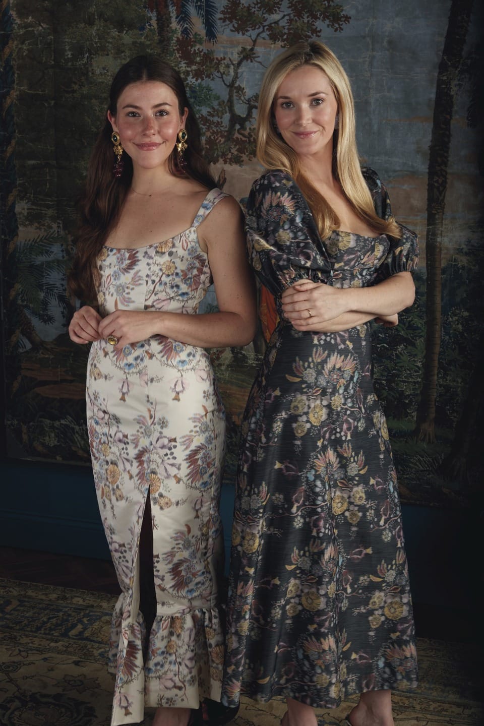Two women in elegant floral dresses standing side by side in front of a decorative backdrop.