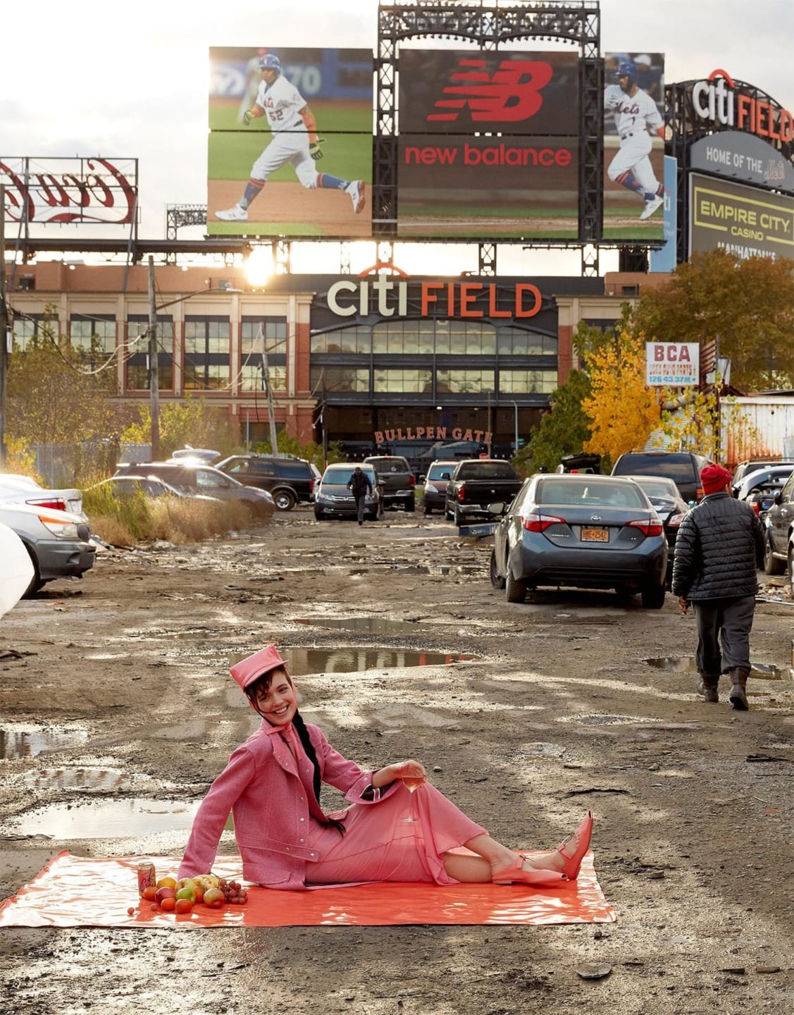 Person in vintage pink outfit having a picnic on a mat in a muddy parking lot near Citi Field with billboards in the background.