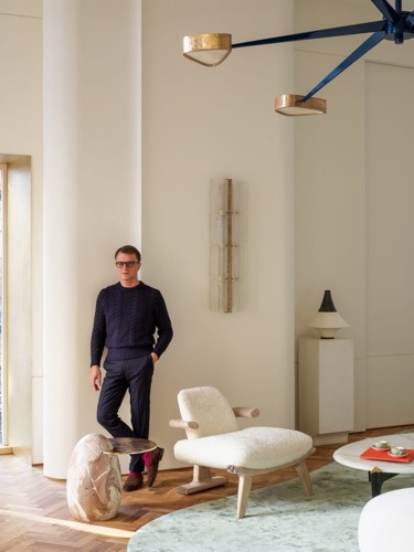 Man in a stylish modern interior with elegant furniture and decor, standing beside a plush chair and a small round table.