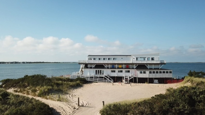 Two-story beachside building surrounded by sand dunes and greenery, with a view of the ocean under a clear blue sky.