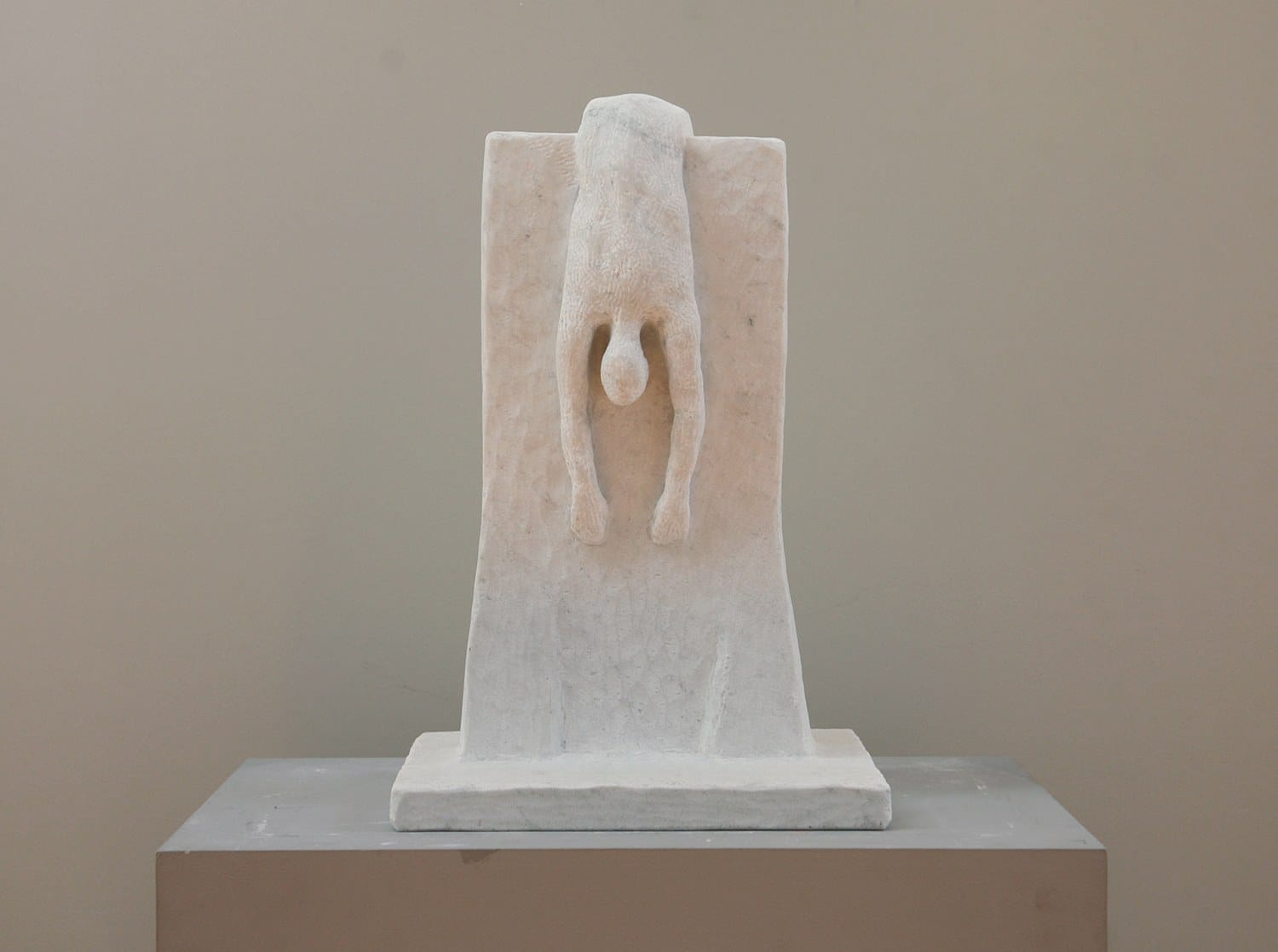 Stone sculpture of a figure mounted on the wall with arms extended downward, displayed against a neutral background.