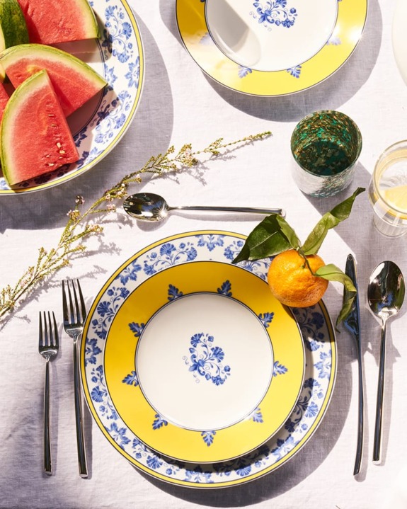 Elegant table setting with floral-patterned plates, silverware, watermelon slices, and an orange on a white tablecloth.