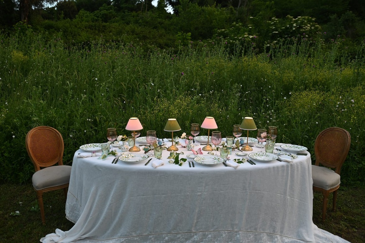 Outdoor dining table set for six with elegant dishware and small lamps, surrounded by green foliage and wildflowers.