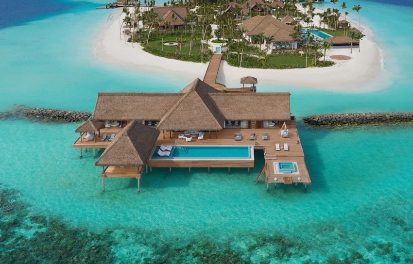 Overwater villa with a private pool and thatched roof on turquoise lagoon near a tropical island with palm trees.