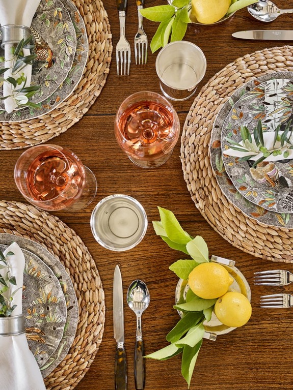Elegant table setting with woven placemats, patterned plates, wine glasses, silverware, lemons, and green leaves on a wooden table.