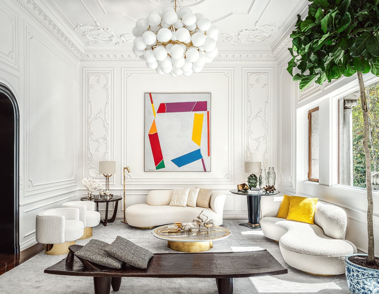 Modern living room with white furniture, abstract wall art, ornate ceiling, large windows, and a lush indoor plant.