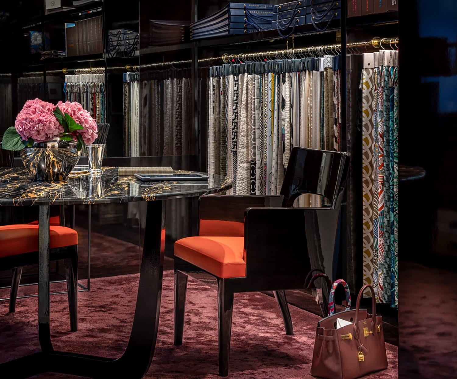 Luxurious interior with a marble table, orange-seat chairs, vibrant pink flowers, and fabric samples in a sophisticated showroom.
