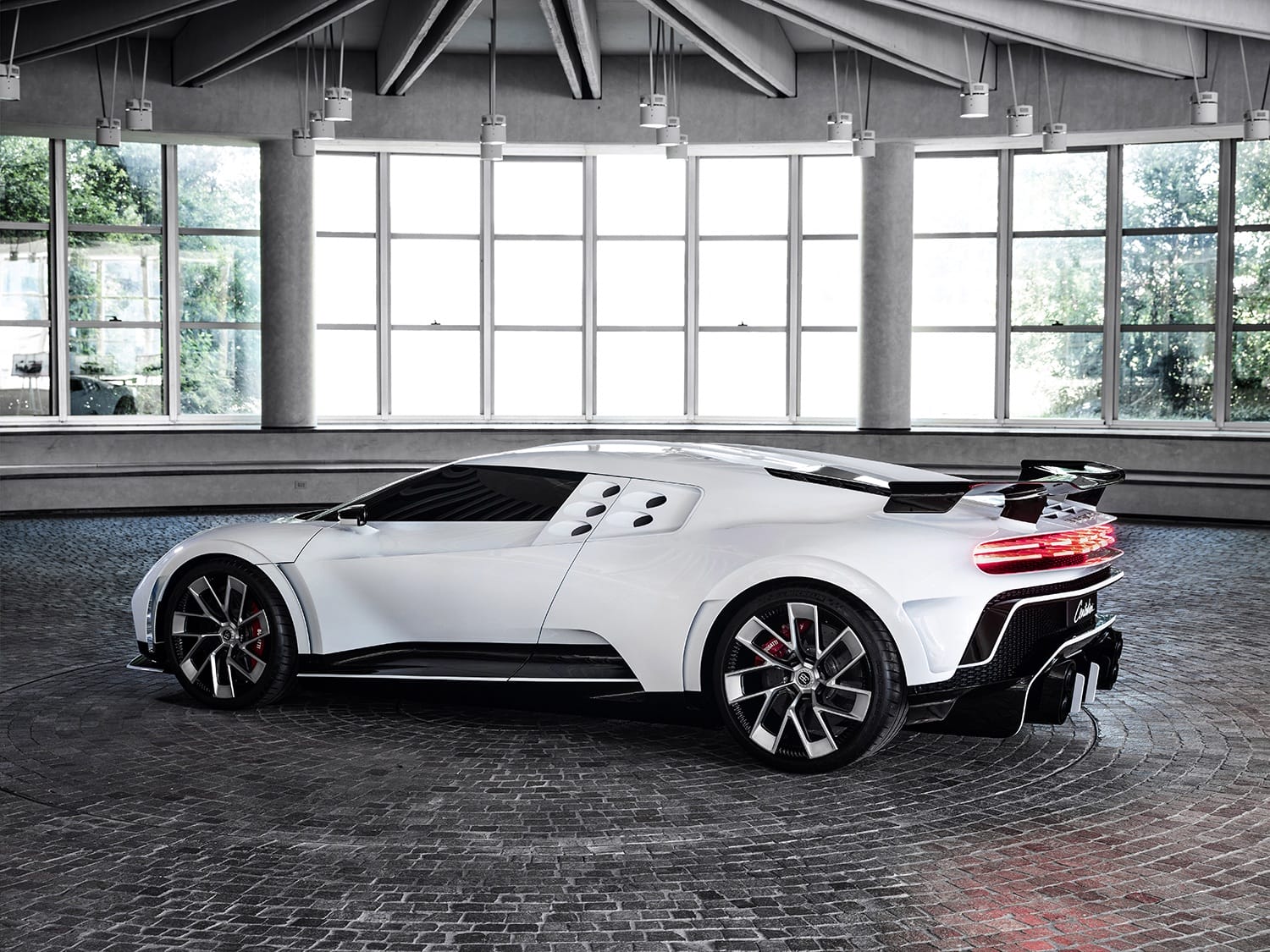 Sleek white sports car with aerodynamic design parked on modern brick floor inside a large room with floor-to-ceiling windows.