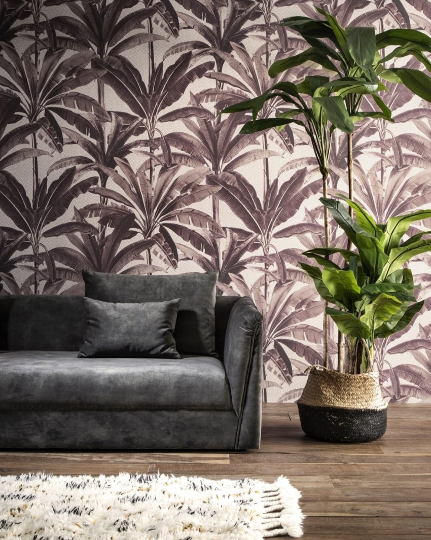 Grey sofa with two pillows in front of tropical leaf-patterned wallpaper, next to a potted plant on a wooden floor.