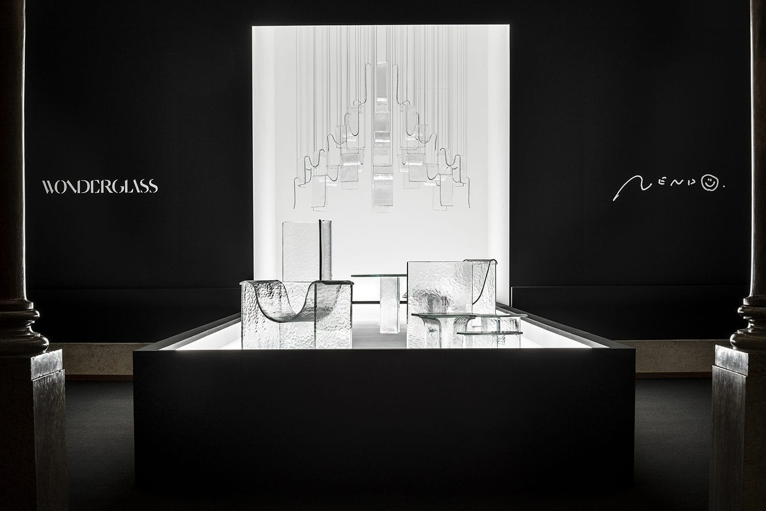 Exhibition of abstract glass sculptures lit dramatically in a dark gallery space with Wonderglass and Nendo logos.