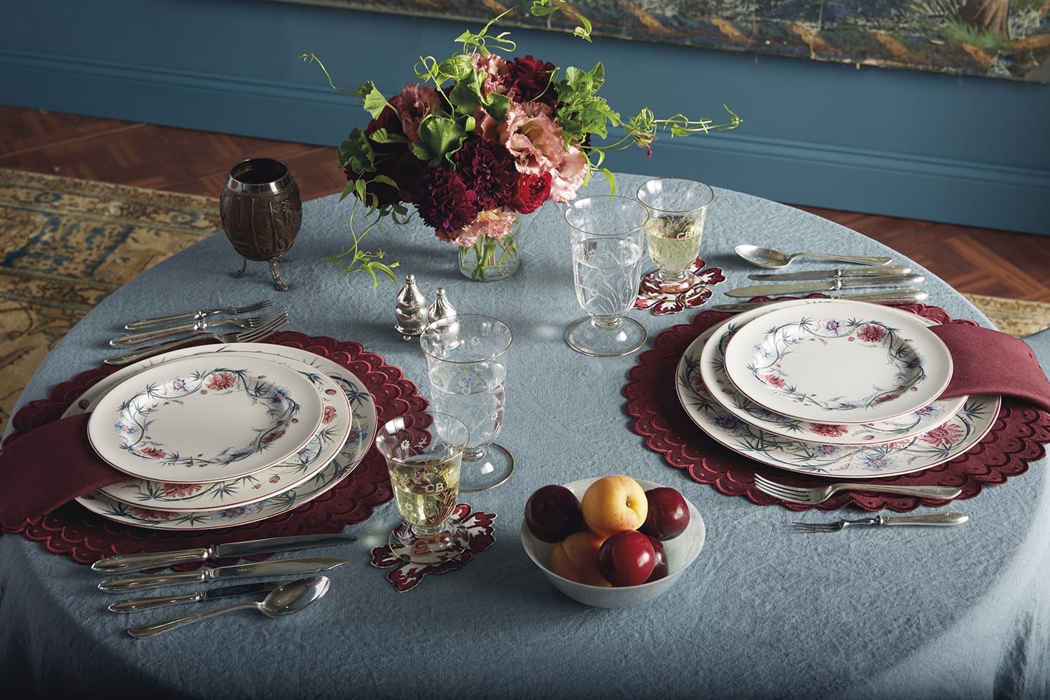 Elegant table setting with floral plates, wine glasses, decorative flowers, and a bowl of fruit on a blue tablecloth.