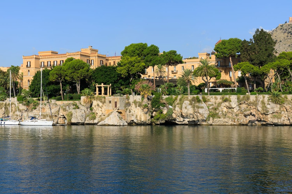 Historic building surrounded by trees on a rocky shoreline, with sailboats on a calm body of water under a clear blue sky.