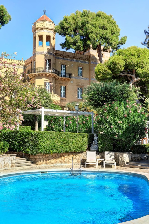 Historic villa with a tower, lush gardens, and a round blue swimming pool in the foreground surrounded by greenery.