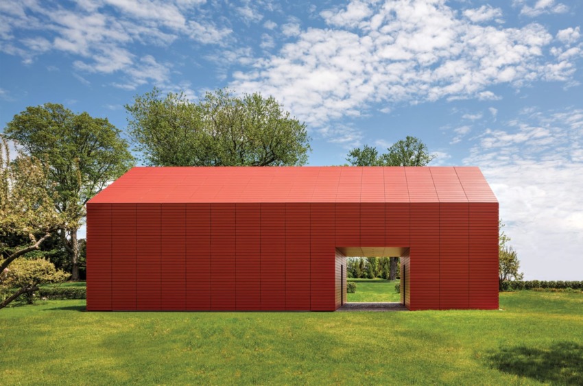 Modern red barn with a large doorway set in a lush green field under a partly cloudy blue sky.