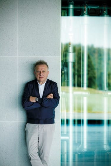 Man standing against a wall with arms crossed, wearing a navy jacket and white shirt, modern building interior in background.