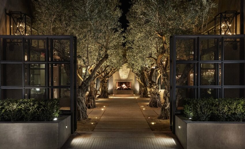 Outdoor courtyard at night with olive trees and glowing fireplace, surrounded by stylish black metal partitions.