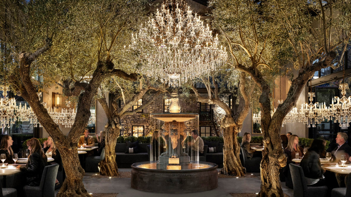 Elegant restaurant interior with chandeliers, trees, and a central fountain, surrounded by people dining.