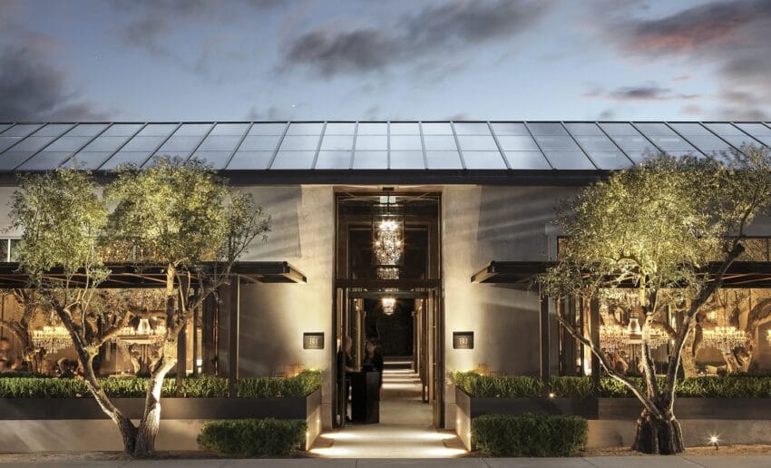Elegant restaurant exterior with modern architecture, glass facade, warm lighting, and surrounding greenery during twilight.