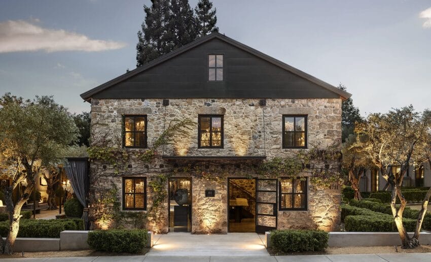 Stone building with ivy, lit windows, surrounded by trees and garden, under a blue sky at dusk.