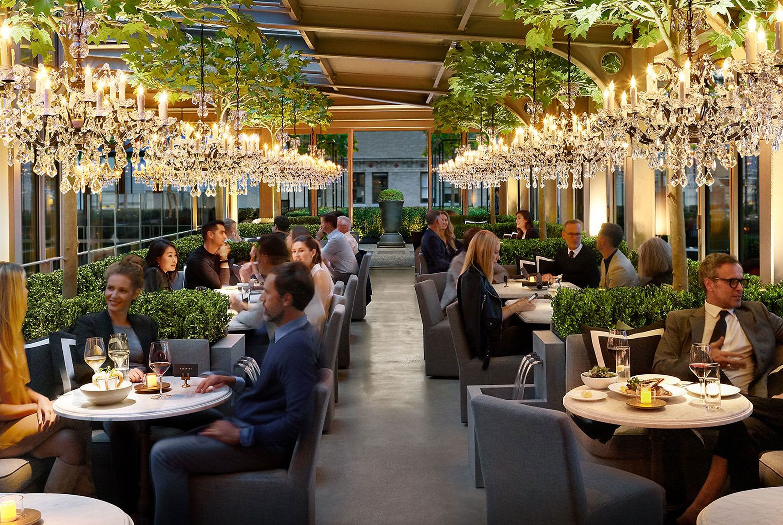 Elegant restaurant with people dining under chandeliers and lush greenery, creating a cozy and sophisticated atmosphere.