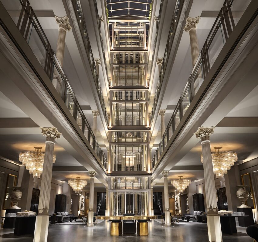 Luxury hotel lobby with grand pillars, chandeliers, and a glass elevator at the center, showcasing elegant architectural design.