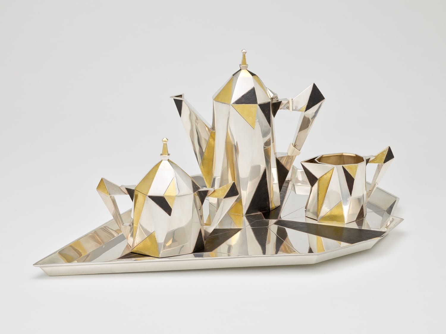 Geometric silver-gold tea set with teapot, sugar bowl, and creamer on an angular tray, Art Deco style.