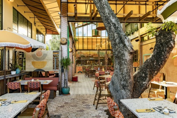 Outdoor cafe with patterned tables, colorful chairs, a large tree, and eclectic decor under a canopy.