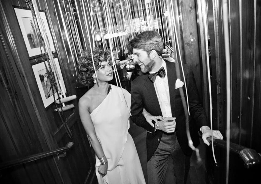 Elegant couple walking through a decorated hallway with streamers, the woman in a white dress and the man in a tuxedo.