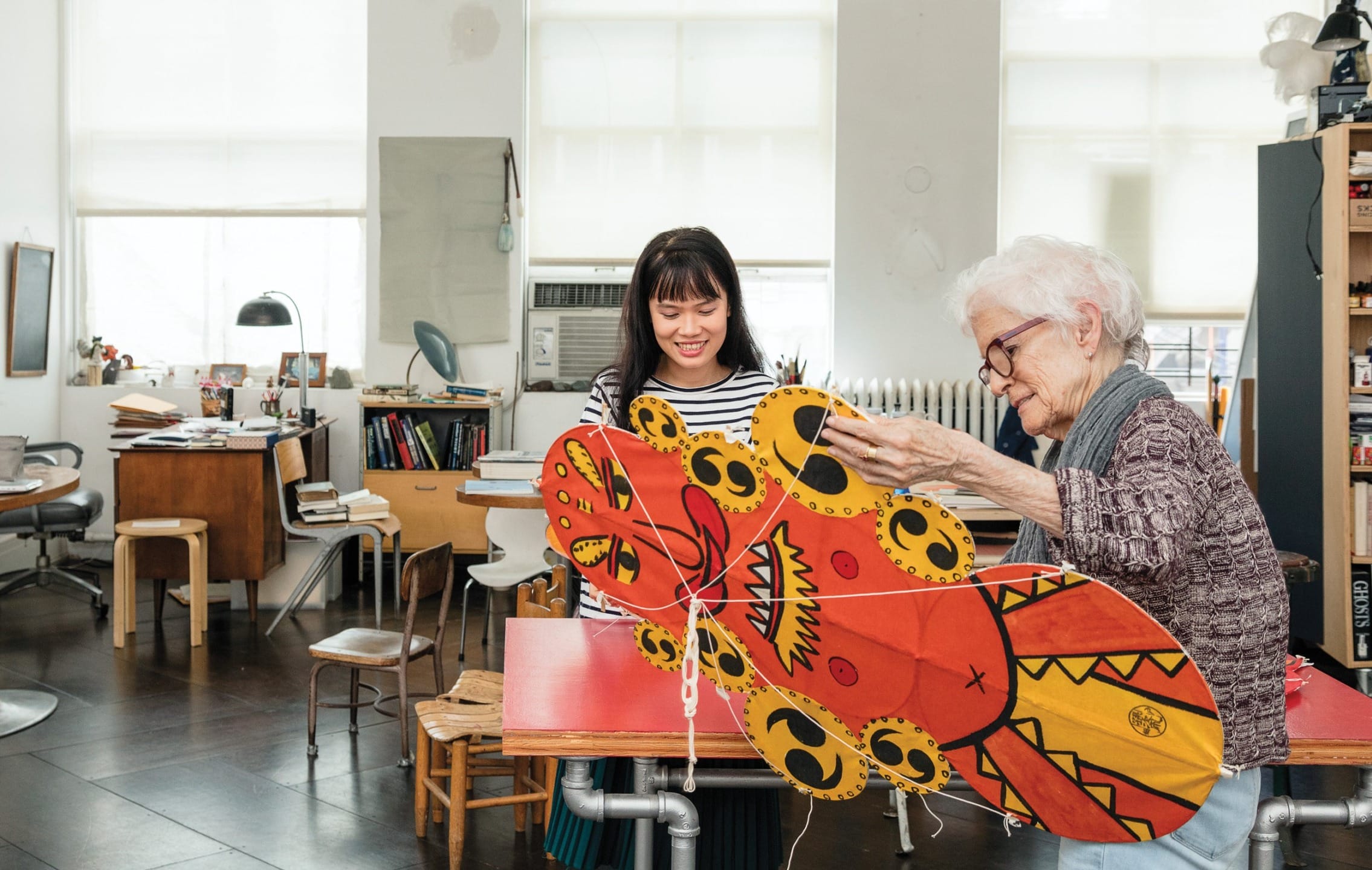 Two women in a studio working on a colorful dragon-shaped kite with vibrant patterns and designs.