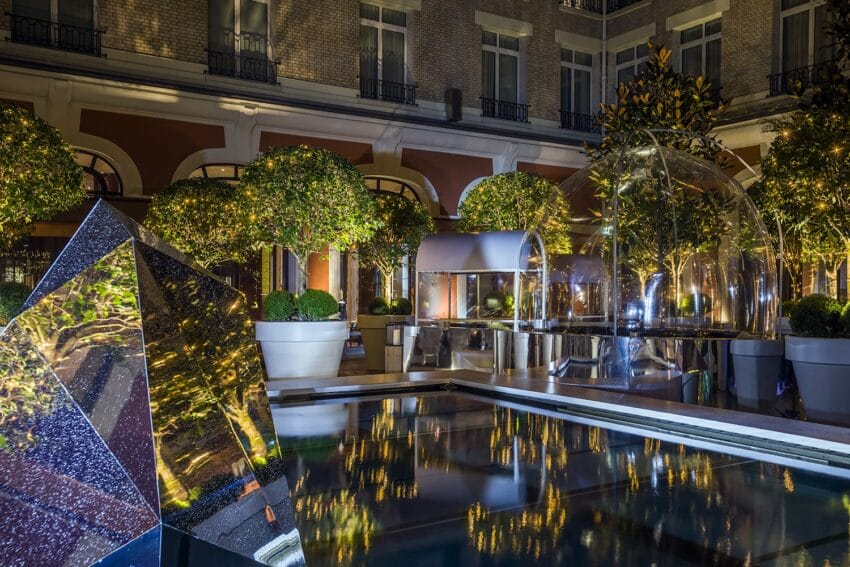 Modern outdoor space with geometric sculptures, water reflections, and illuminated greenery at night, outside a classic building.