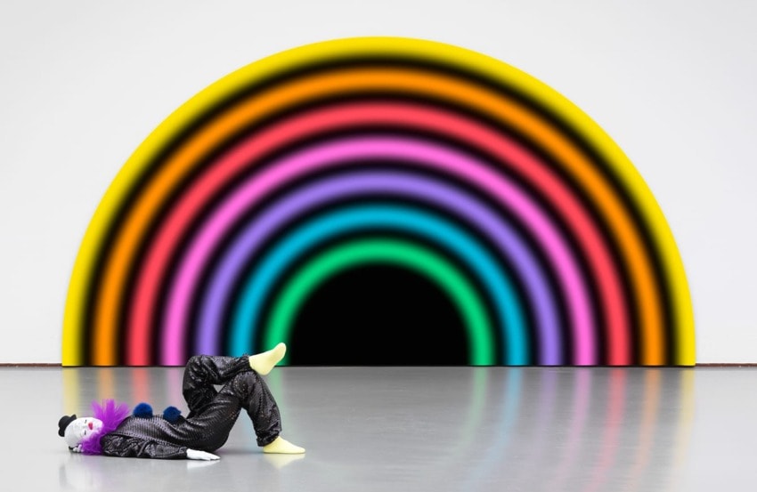 Clown lounging on the floor in front of a large, colorful rainbow mural with bright, vivid concentric arcs.