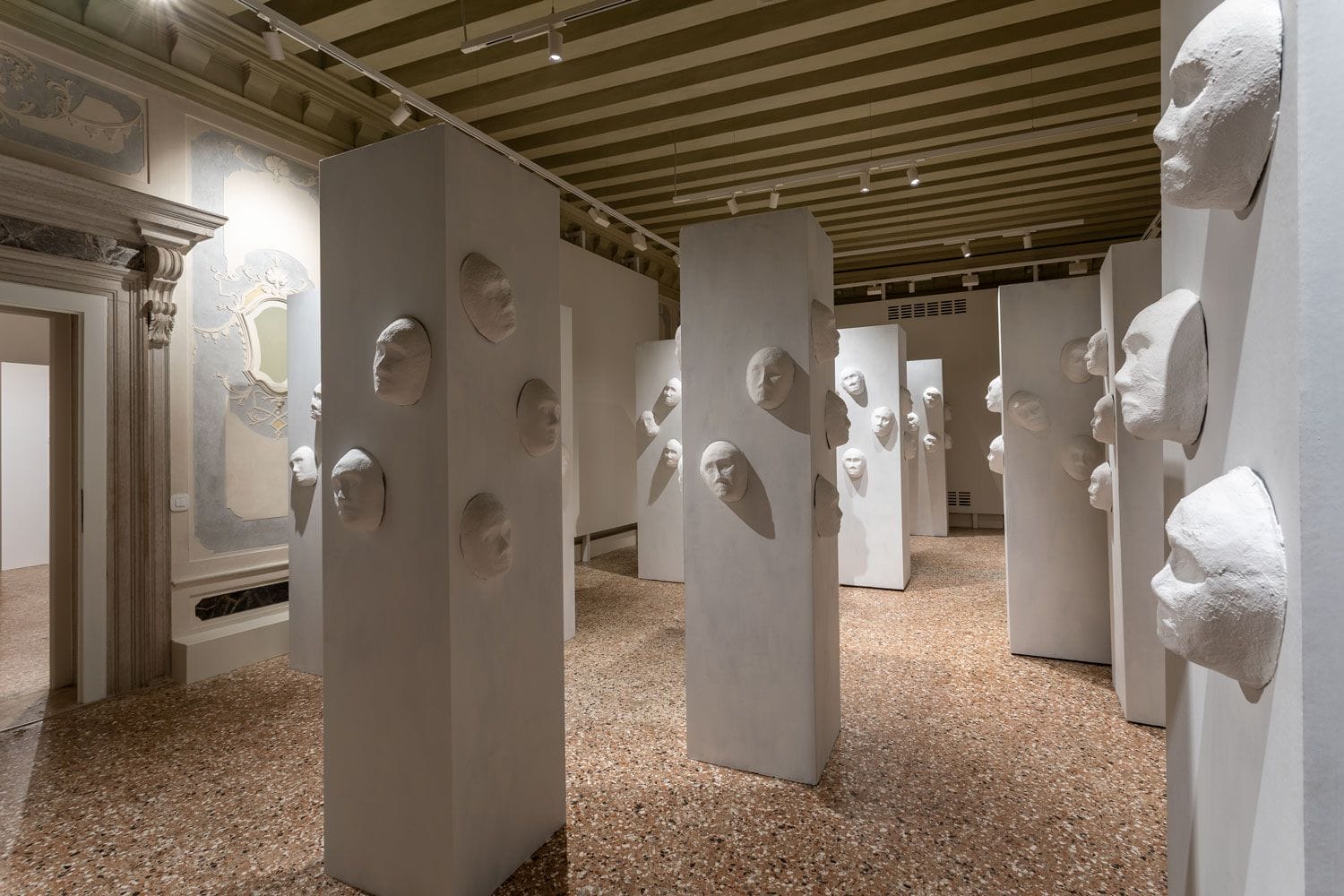 Art installation with white plaster faces mounted on tall pillars in a gallery with ornate walls and wooden ceiling beams.