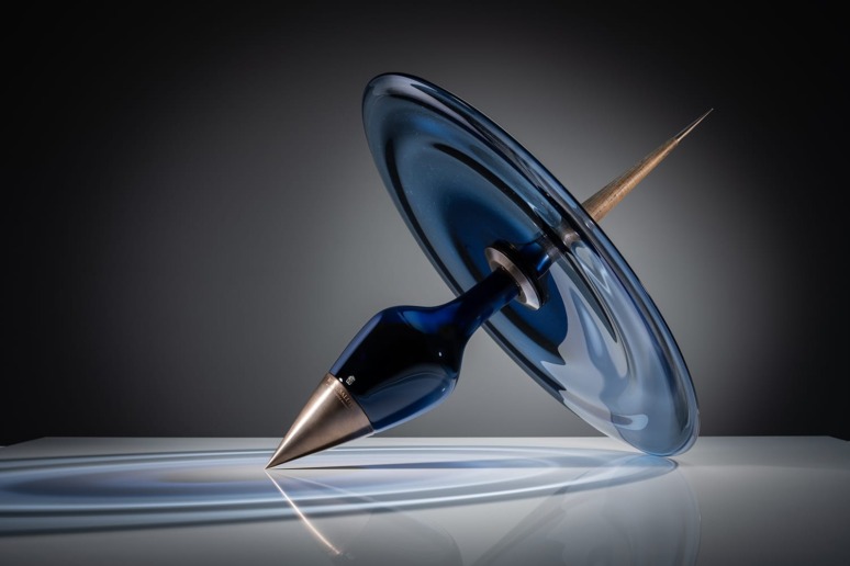 Blue glass spinning top with a metallic tip spinning on a reflective surface against a dark background.