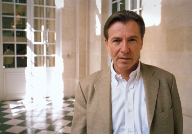 Man in a beige blazer and white shirt standing in a sunlit hallway with checkered floors.