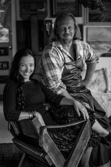 Two people smiling in a cozy, artistic studio setting with paintings in the background.