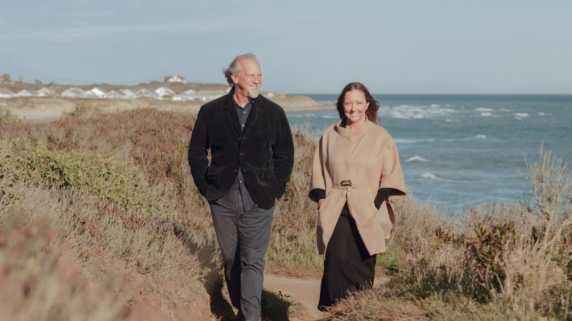 A man and a woman walking on a coastal path, with the ocean and distant cliffs in the background.