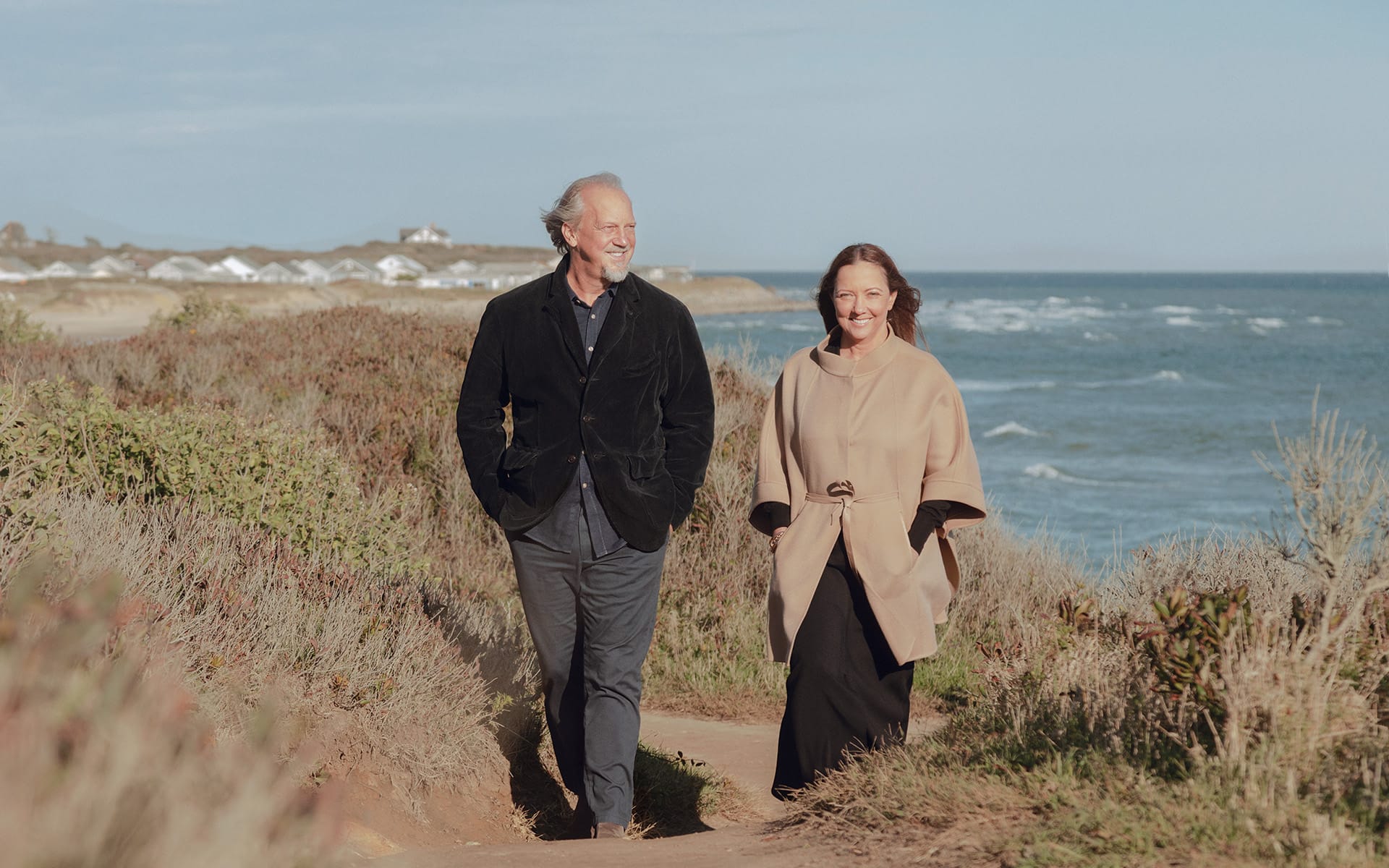 A man and a woman walking on a coastal path, with the ocean and distant cliffs in the background.