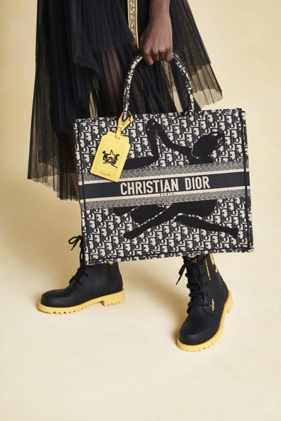 Person holding a Christian Dior tote bag with a black and yellow color scheme, wearing matching boots on a beige background.