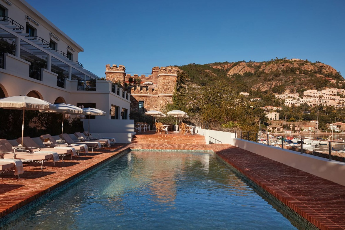 Outdoor hotel pool with lounge chairs and umbrellas overlooking scenic hills and buildings under a clear blue sky.