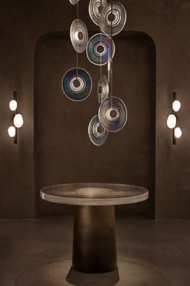 Modern art installation with circular glass discs and elegant lighting above a round, minimalist table in a dimly lit room.