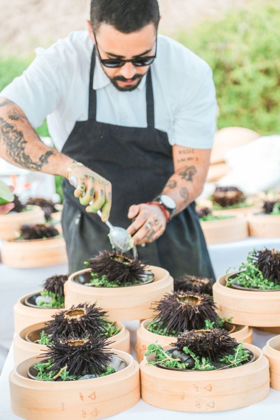 Chef preparing sea urchin dishes on wooden plates outdoors, wearing apron and sunglasses, with tattooed arms.
