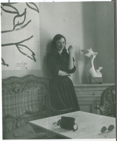 Woman in a dark dress poses near wicker chairs and abstract art in a stylish room with a sculpture on the table.