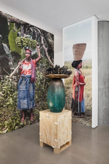 Art installation with large photographs of people, green vase on wooden box, concrete wall in gallery setting.