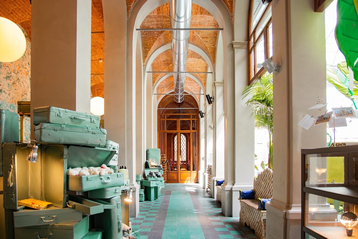 Colorful, vintage corridor with arched ceiling, teal luggage, and natural light streaming through large windows.
