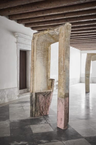 Stone gateway sculpture displayed in a minimalist gallery space with wooden beams and large windows