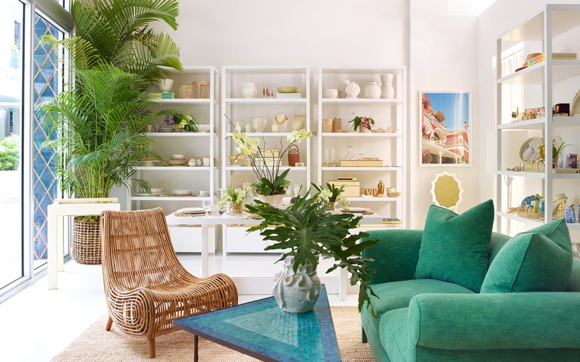 Modern living room with green sofa, wicker chair, plants, and white shelves displaying decorative items. Bright and airy space.