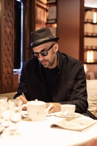 Man in a black hat and sunglasses writing at a table in a cozy, dimly lit restaurant with a teapot and cups nearby.